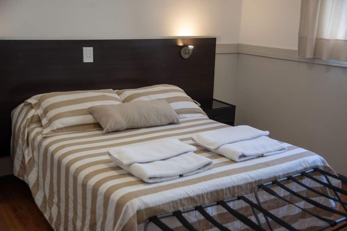 A neatly made bed with striped bedding and a headboard, a bedside table with a lamp, and a window with curtains.
