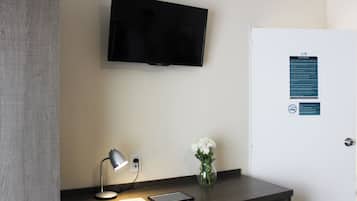 Una habitación de hotel con televisión de pantalla plana en la pared, un escritorio con lámpara, un jarrón con flores y una puerta con cartel.
