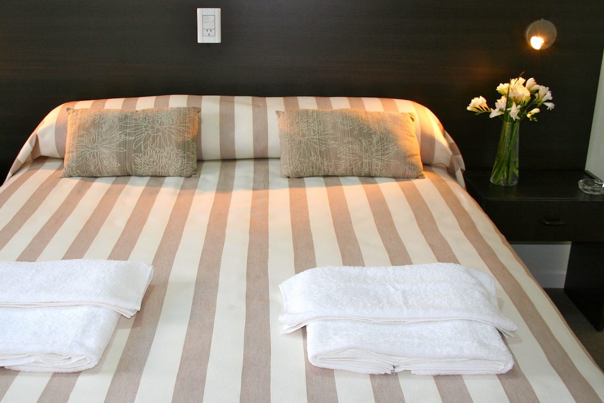 A hotel room with a striped bedspread, two pillows, a bedside table with a phone, a vase with flowers, and a wall socket.
