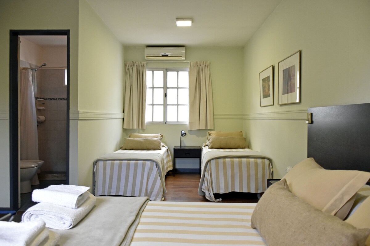 Habitación de hotel con dos camas individuales, ducha, ventana con cortinas y aire acondicionado instalado en la pared.