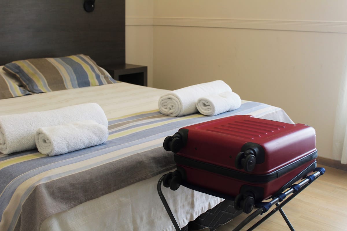 A bed with striped bedding and rolled white towels, a red suitcase on a luggage cart, and a nightstand with a lamp.