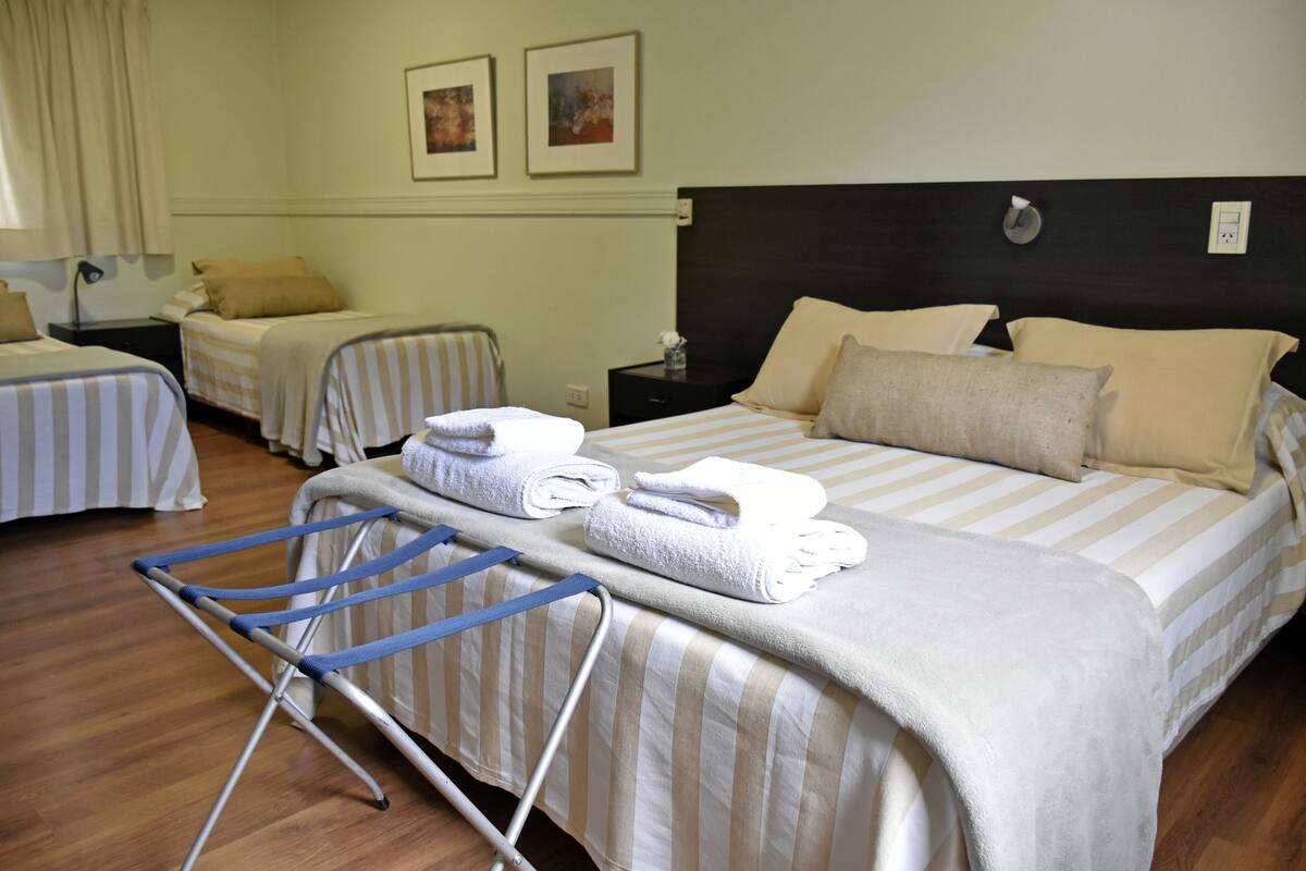 A hotel room with two single beds, a large bed, a blue ironing board, and a wall-mounted power outlet.