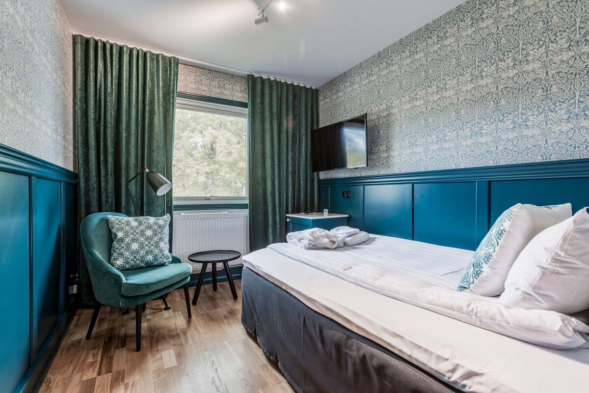 A hotel room with a large bed, a chair, a small table, a TV, and a window with curtains.