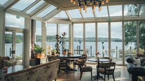 A spacious room with large windows overlooking a lake, furnished with a round table, chairs, and a sofa. - Hällsnäs (Mölnlycke)