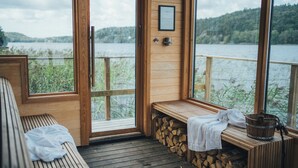 A wooden deck with a bench and a fireplace, overlooking a lake with a forested hill in the background. - Hällsnäs (Mölnlycke)