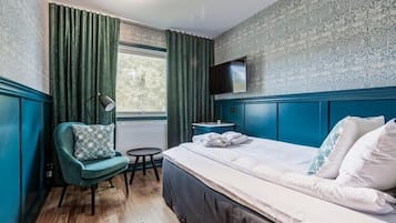 Ett hotellrum med en stor säng, en stol, ett litet bord, en tv som är monterad på väggen och ett fönster med gardiner.