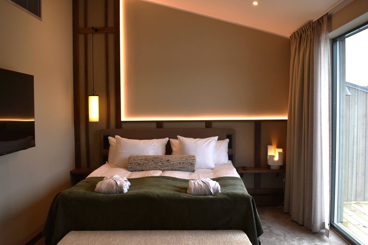 A neatly made bed with a green blanket, white pillows, and a decorative pillow. There are two bedside lamps and a flat-screen TV mounted on the wall.