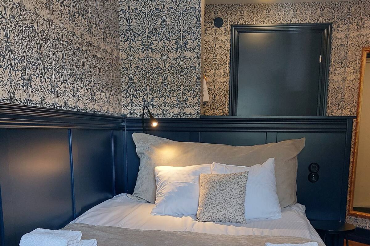 A neatly made bed with white linens and a folded towel on the side, a bedside table with a lamp, and a patterned wallpaper.