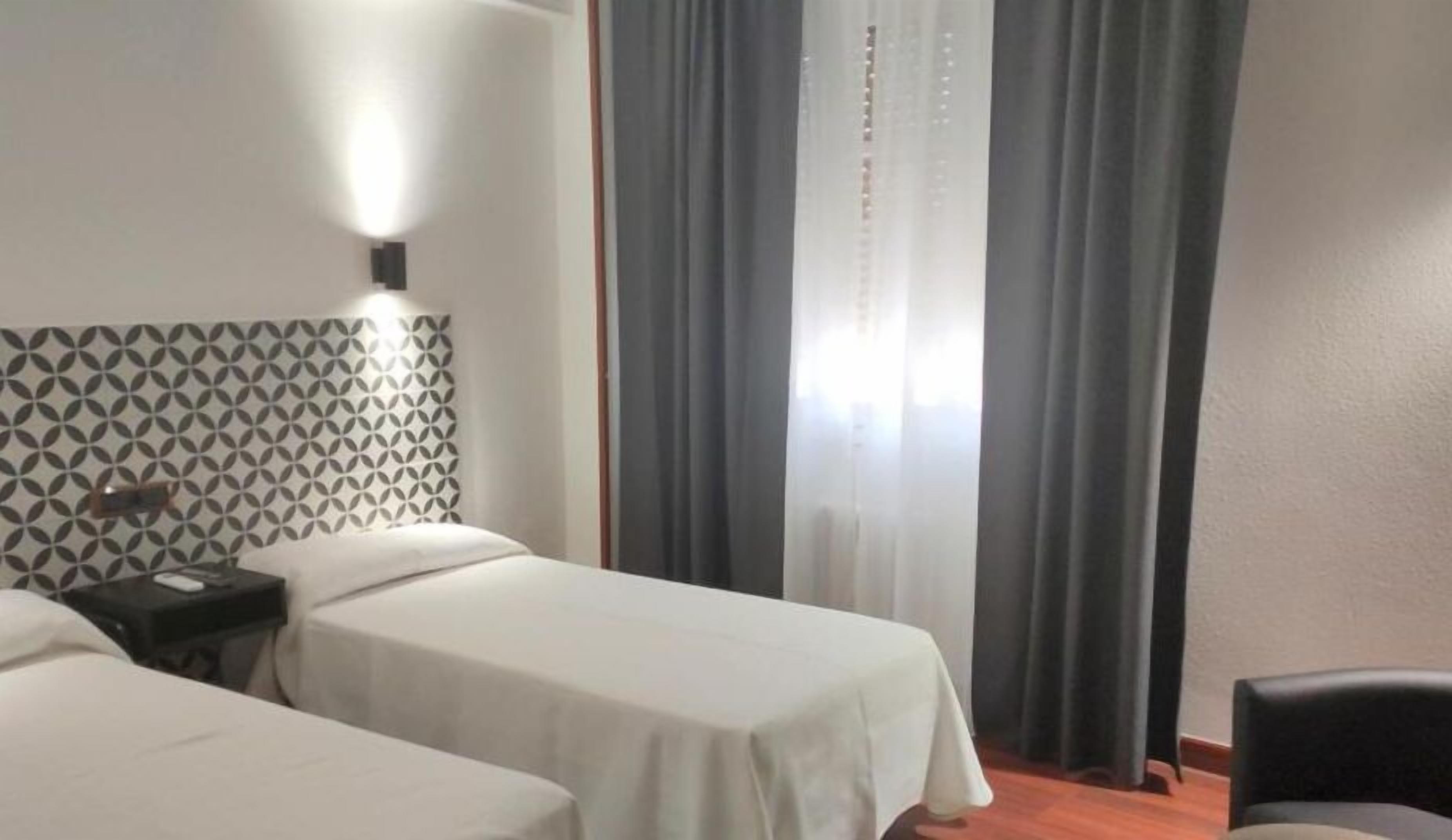A hotel room with two single beds, a patterned headboard, a bedside table with a lamp, and a window with curtains.