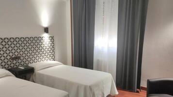 A hotel room with two single beds, a patterned headboard, a bedside table with a lamp, and a window with curtains.