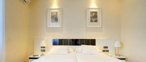 Una cama doble con ropa de cama blanca, dos mesitas de noche con lámparas y dos cuadros en la pared.