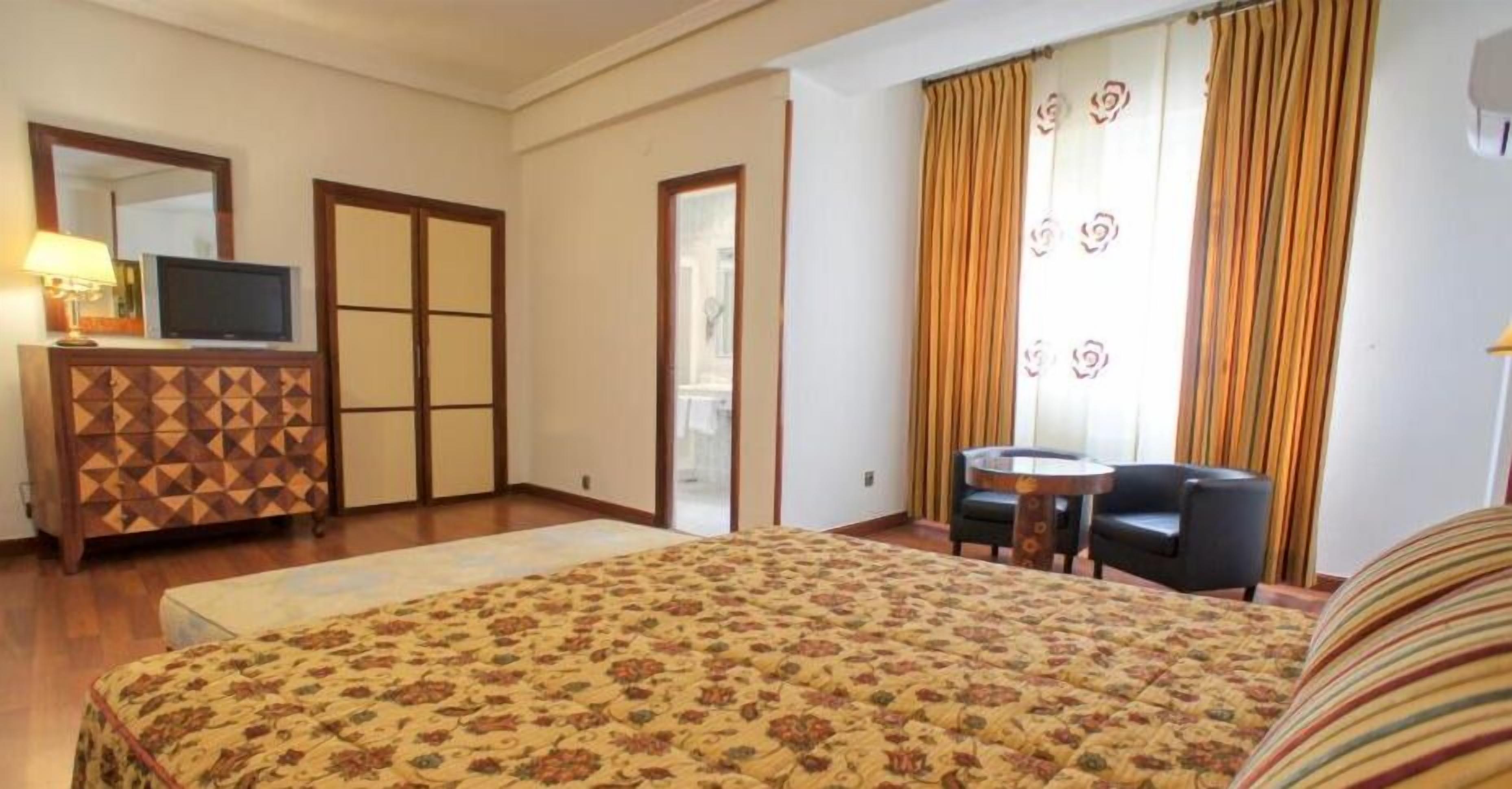 Habitación de hotel con cama, escritorio, silla, televisor, cómoda y ventana con cortinas.