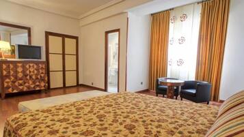 Habitación de hotel con cama, escritorio, silla, televisor, cómoda y ventana con cortinas.