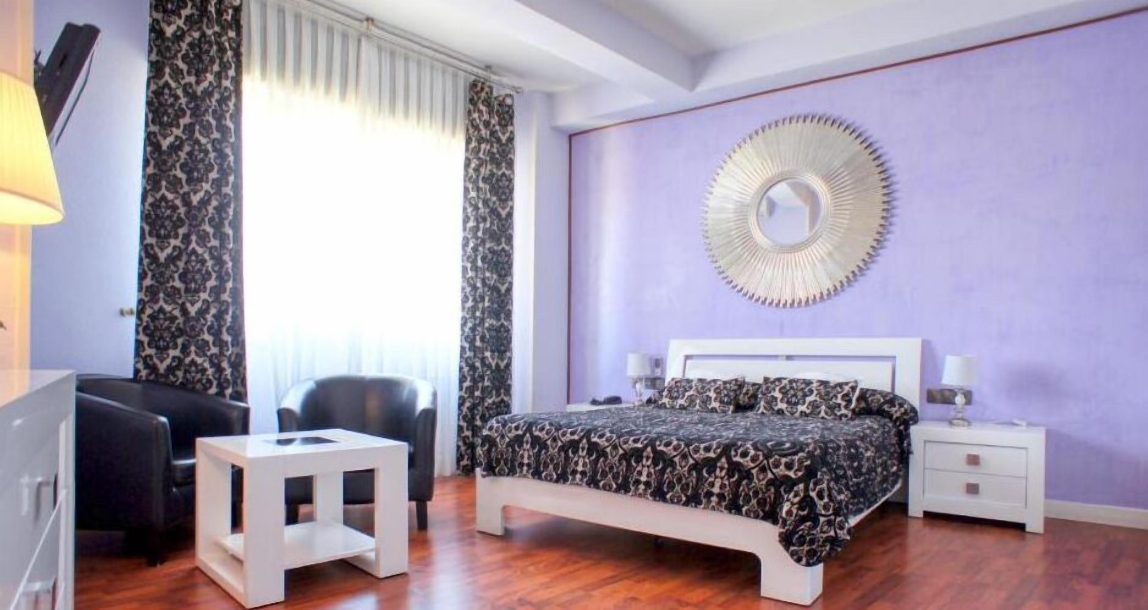 A bedroom with a bed, a small table, a chair, a nightstand, and a mirror on the wall.