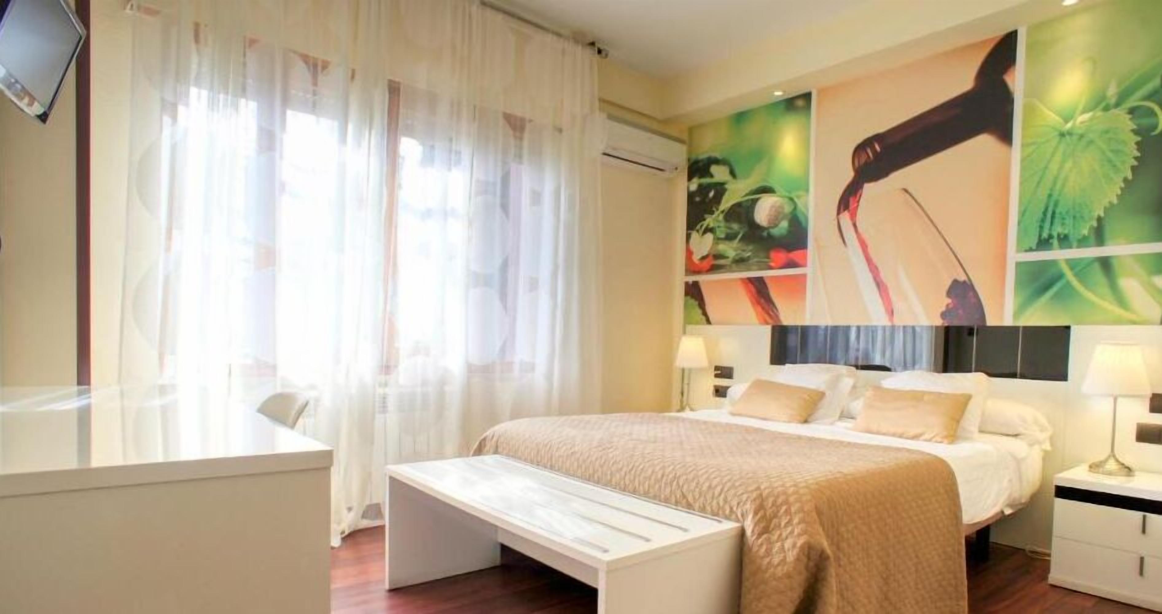 A modern hotel room with a bed, bedside tables, a desk, and a large window with sheer curtains.