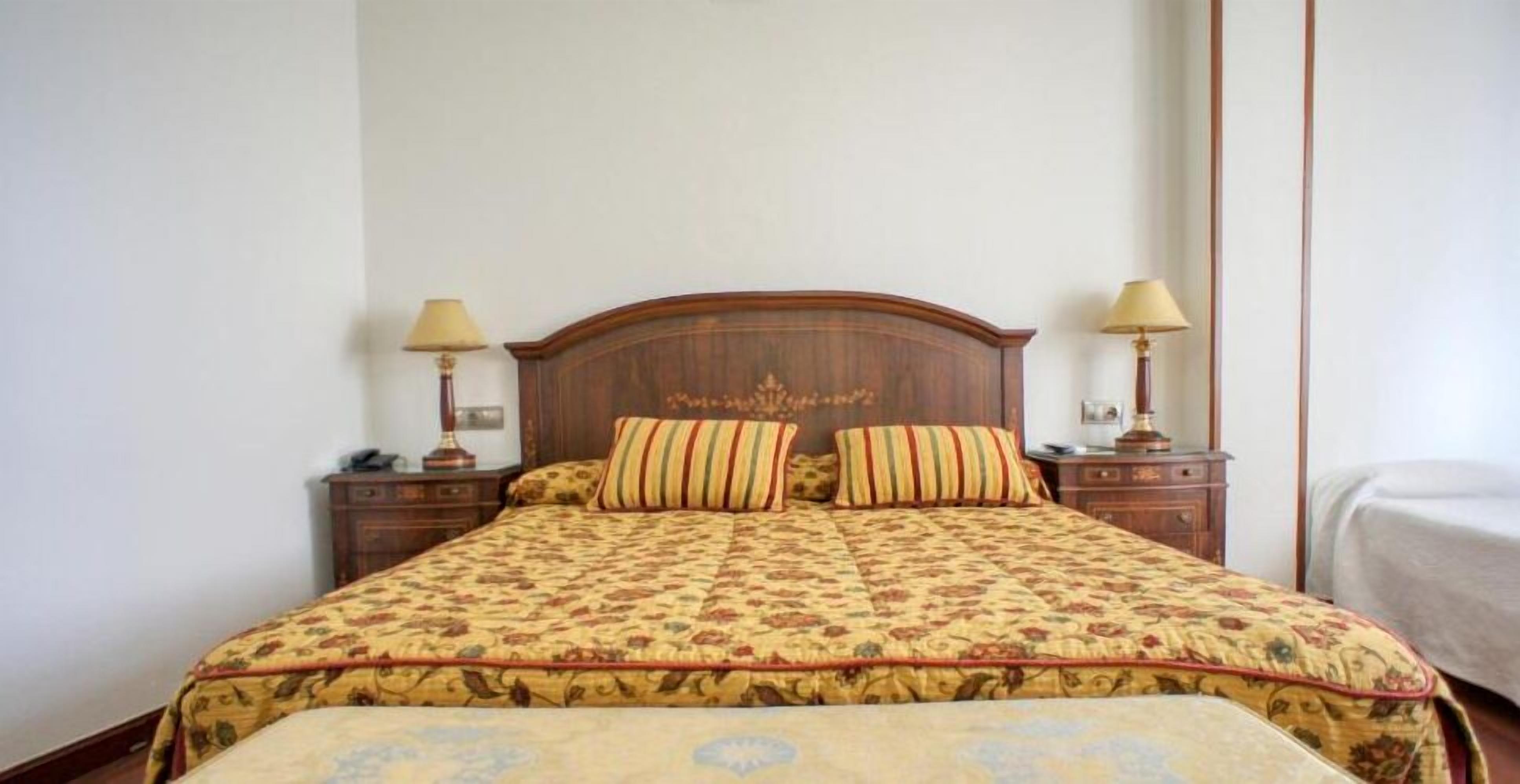 Una cama bien tendida con una colcha de motivos florales y dos almohadas a juego, flanqueada por mesitas de noche de madera con lámparas.