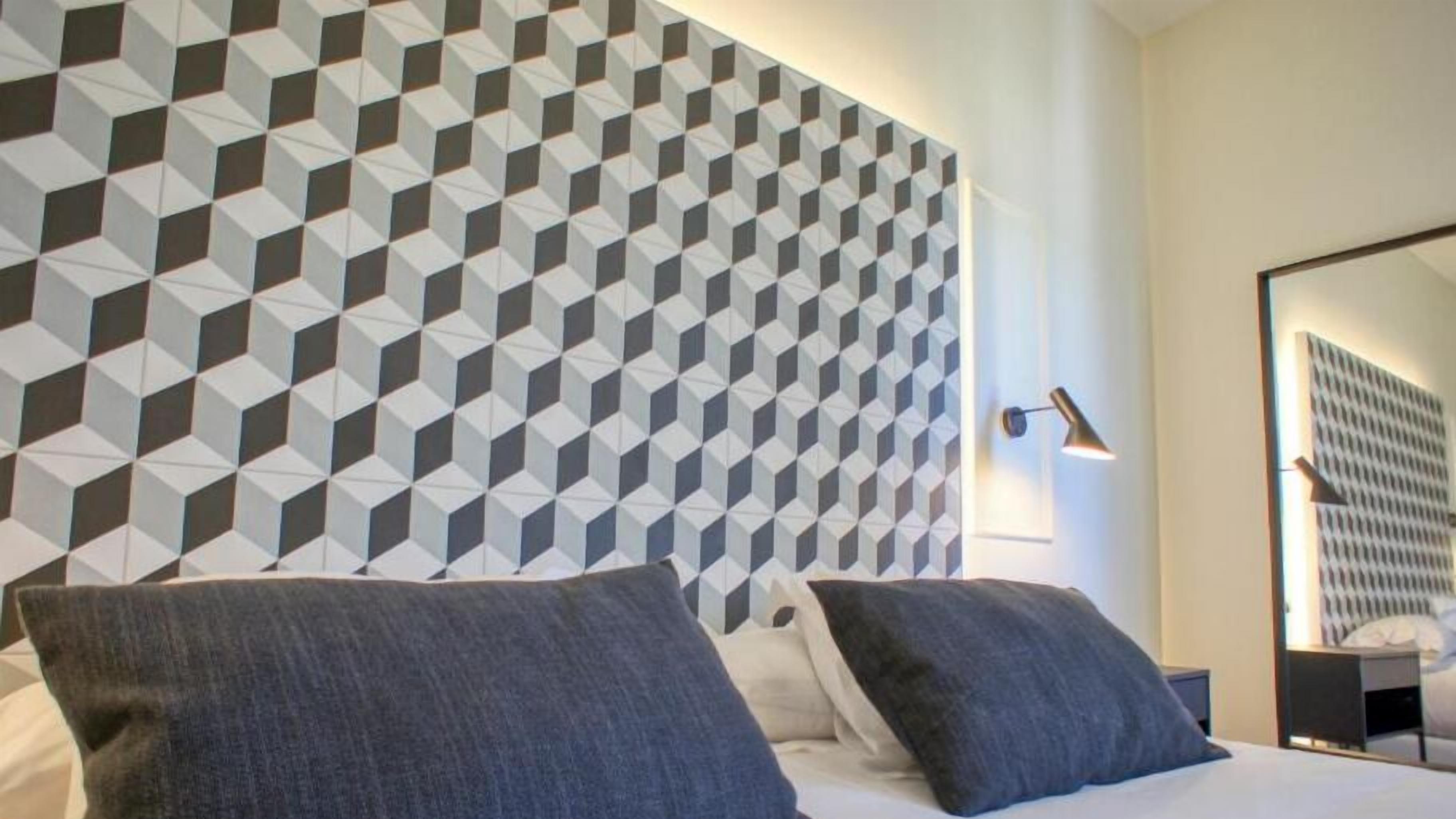 A hotel room with a geometric patterned wall, a bed with two pillows, a nightstand, a lamp, and a large mirror.