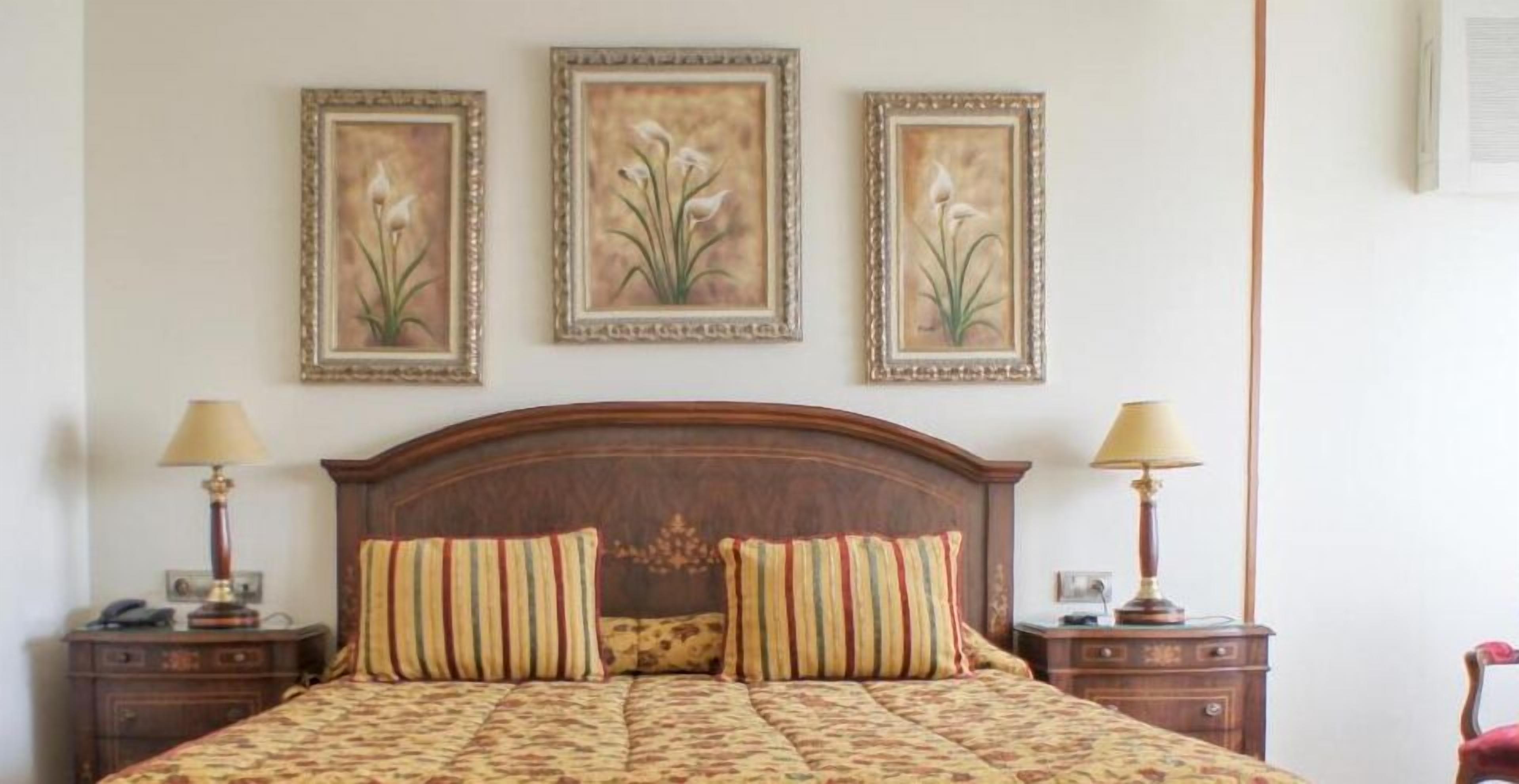 A hotel room with a wooden headboard, two bedside tables with lamps, and three framed floral paintings on the wall.