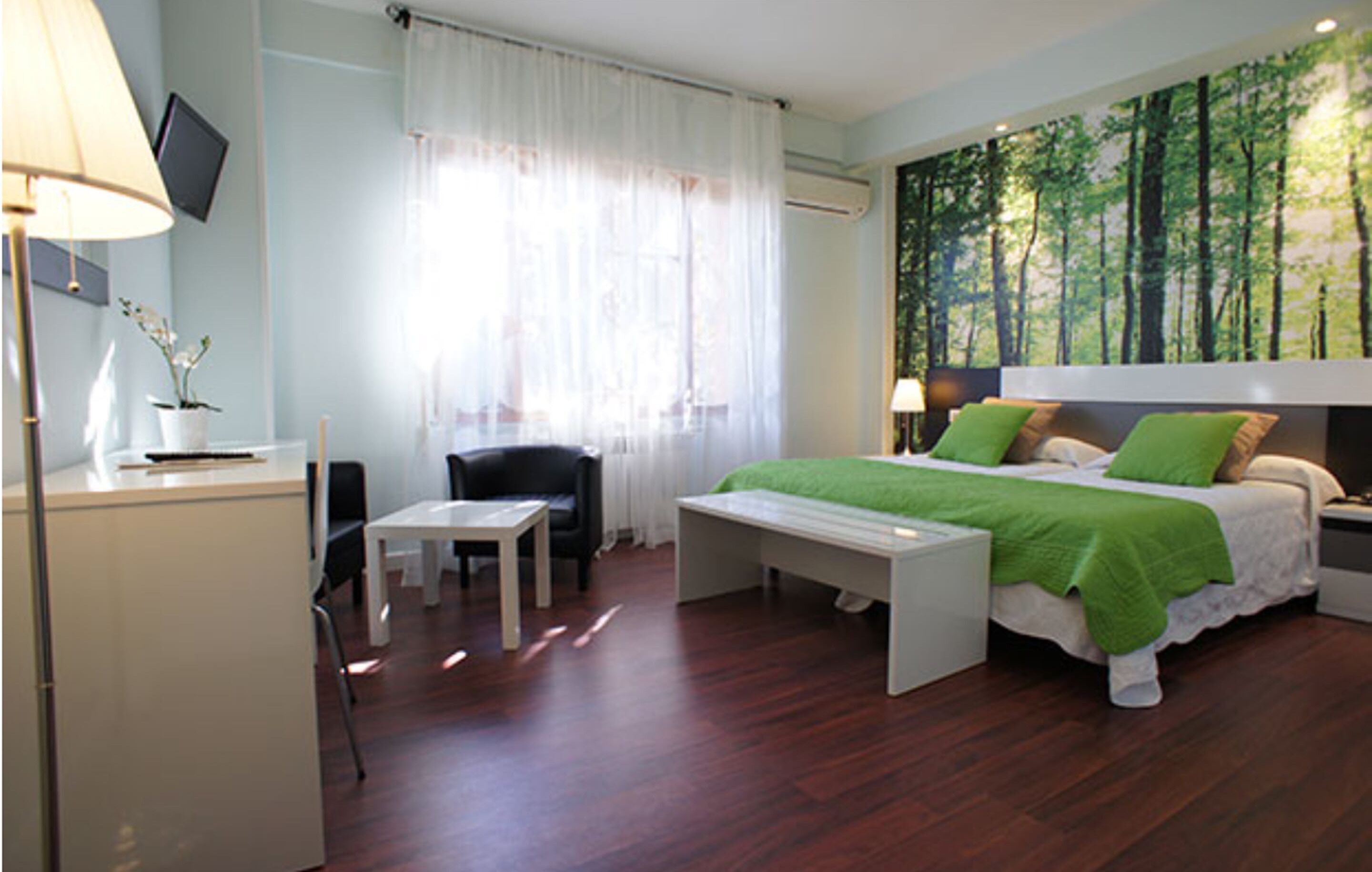 A bedroom with a bed, bedside table, chair, desk, and a TV on the wall.
