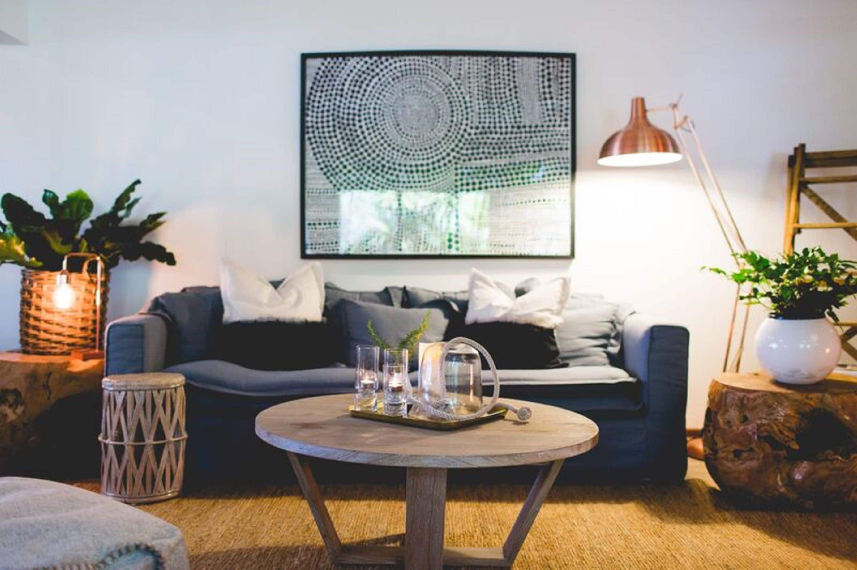 A cozy living room with a round coffee table, a blue sofa with cushions, a large abstract painting, and a floor lamp.