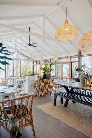 A spacious dining area with a high ceiling, large windows, and a variety of seating options including wicker chairs and a wooden bench.