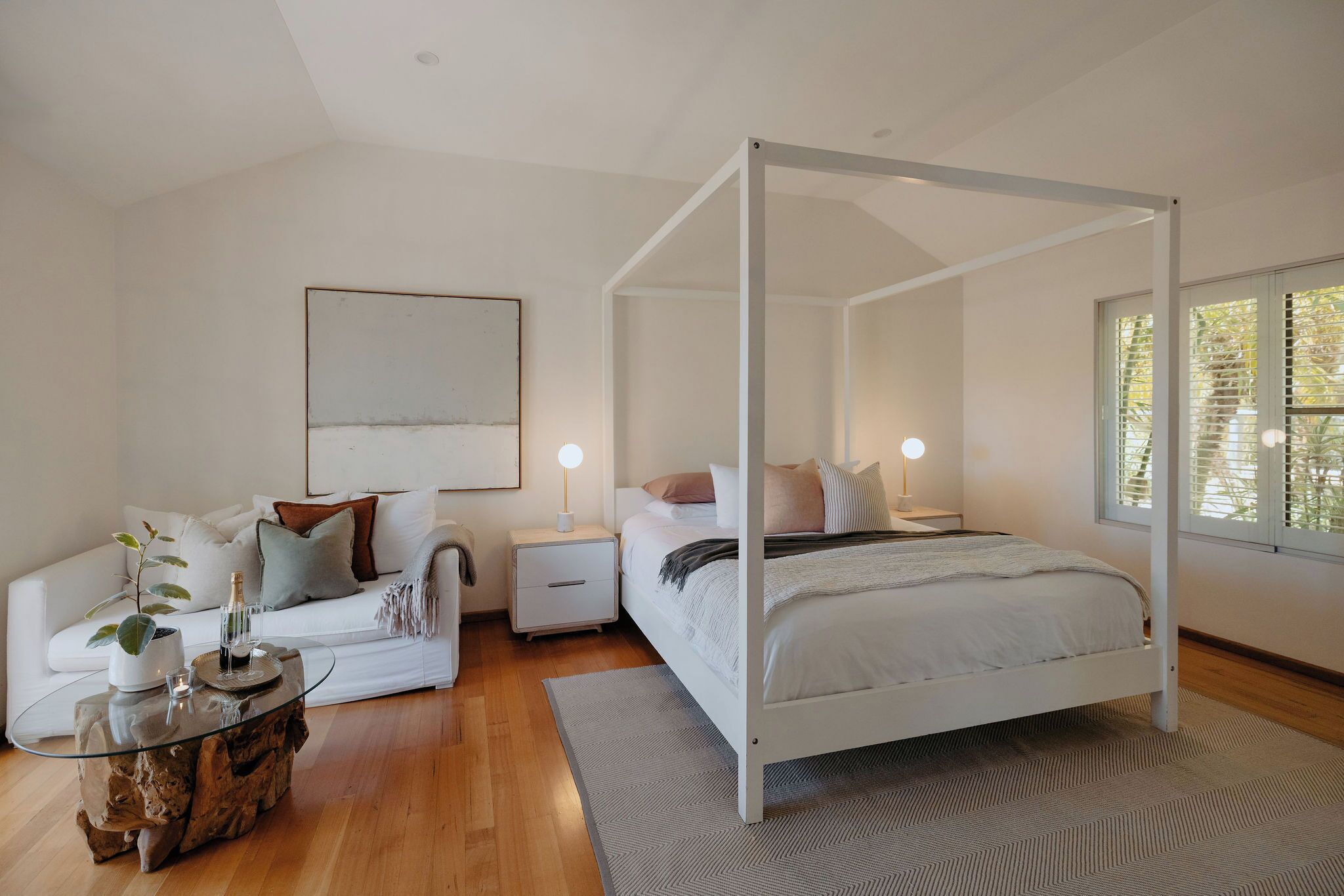 Une chambre à coucher avec un lit à baldaquin, un canapé blanc, une table de chevet avec une lampe et une grande fenêtre avec des stores.