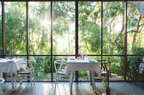 A dining area with large windows offering a view of a lush garden. Tables are set with white tablecloths and chairs. - Yala Yallingup Retreat and Spa (Quedjinup)