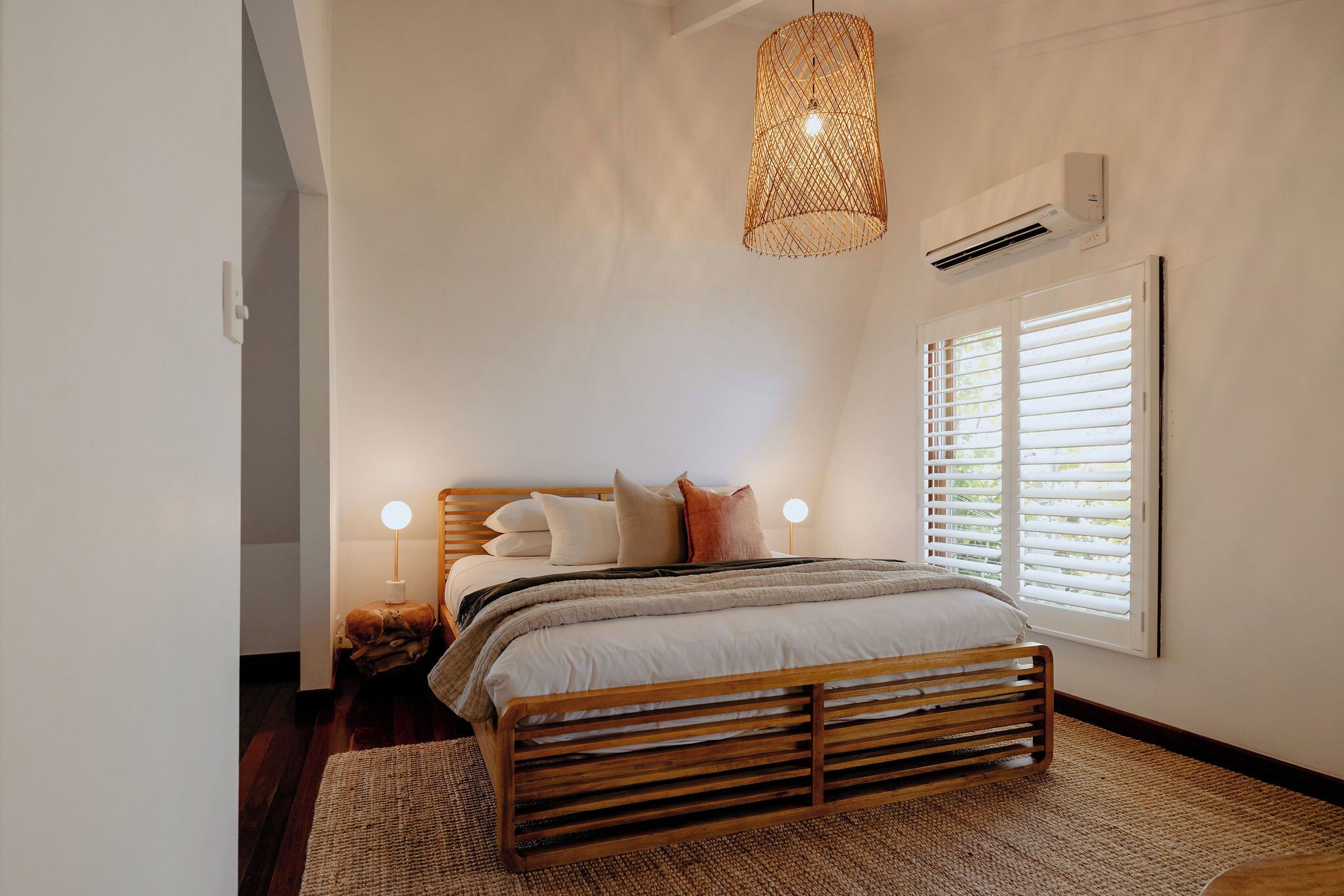 A bedroom with a wooden bed, a woven light fixture, a bedside lamp, and a window with blinds.