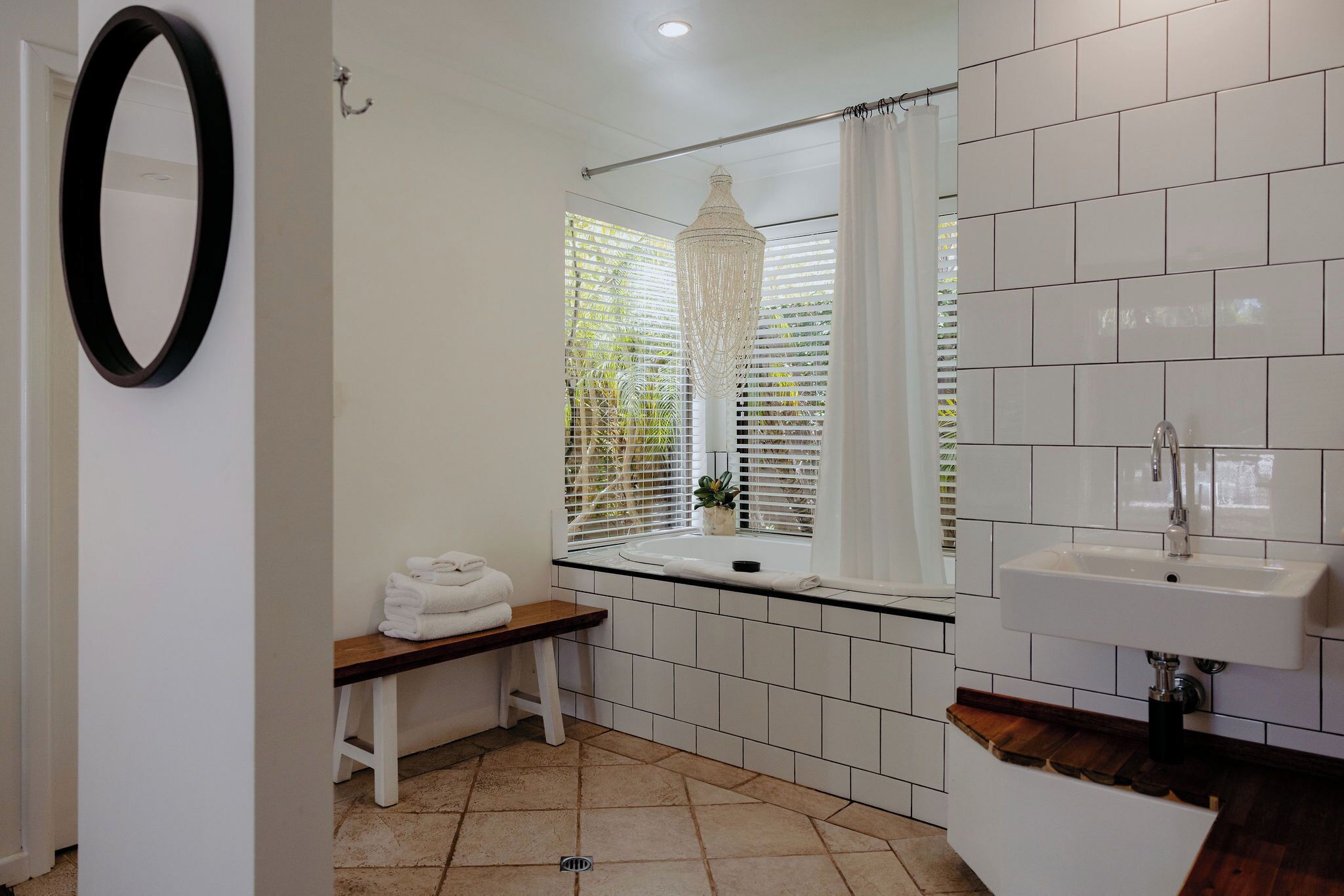 A bathroom with white tiles, a sink, a bench with towels, and a window with blinds.