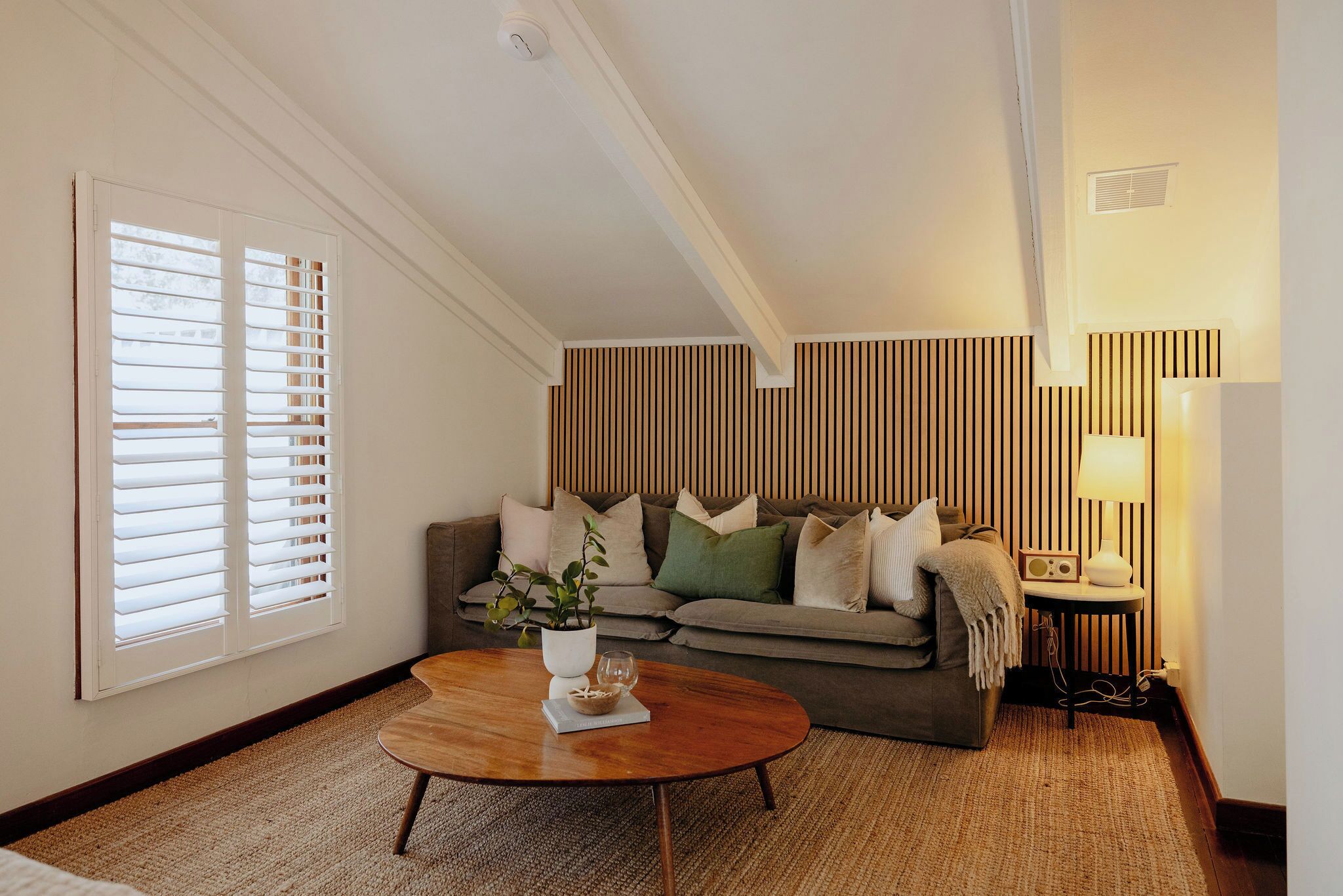 Una sala de estar acogedora con una mesa de centro de madera, un sofá con cojines, una mesita de noche con lámpara y una ventana con persianas.