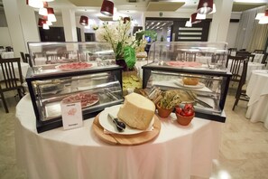 Una mesa de buffet con diversos alimentos, incluyendo un bloque grande de queso, una cesta de pan y un plato con salchichas etiquetado "SALAME Sausages".