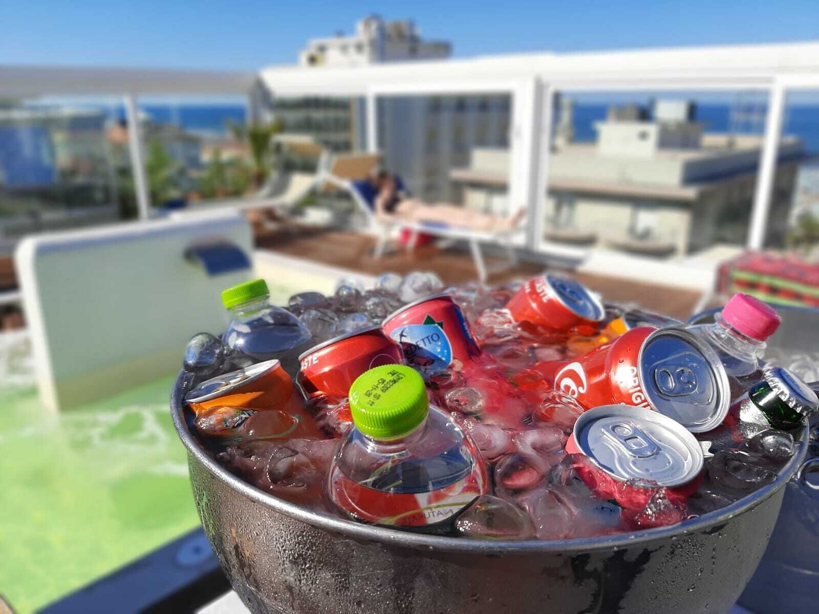 Ein Eimer voller Eis und verschiedener Getränkedosen und -flaschen auf einer Dachterrassen mit Blick auf Gebäude und das Meer.