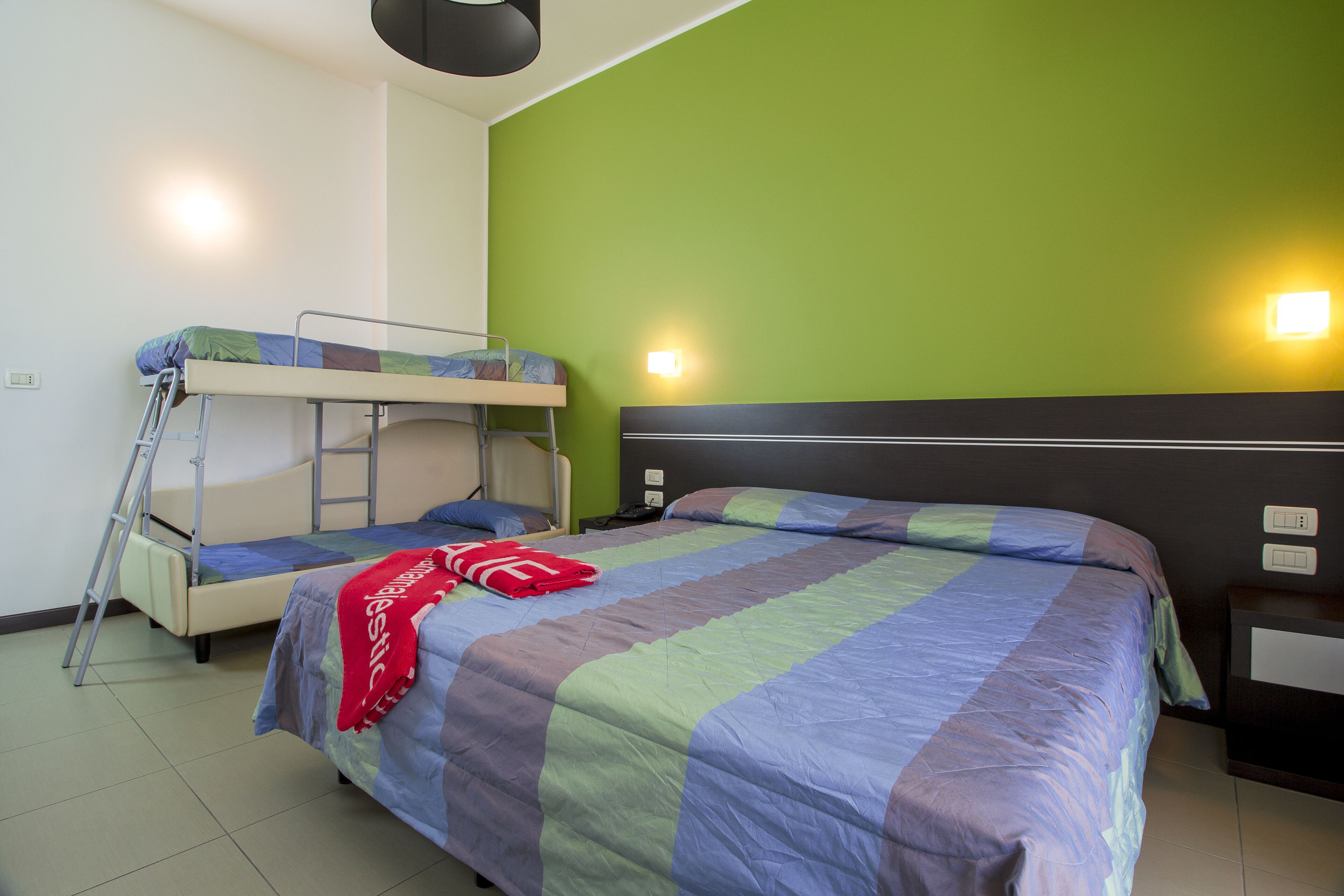 A room with bunk beds, a bed with a striped blanket, a ladder, bedside tables, and wall-mounted lights.