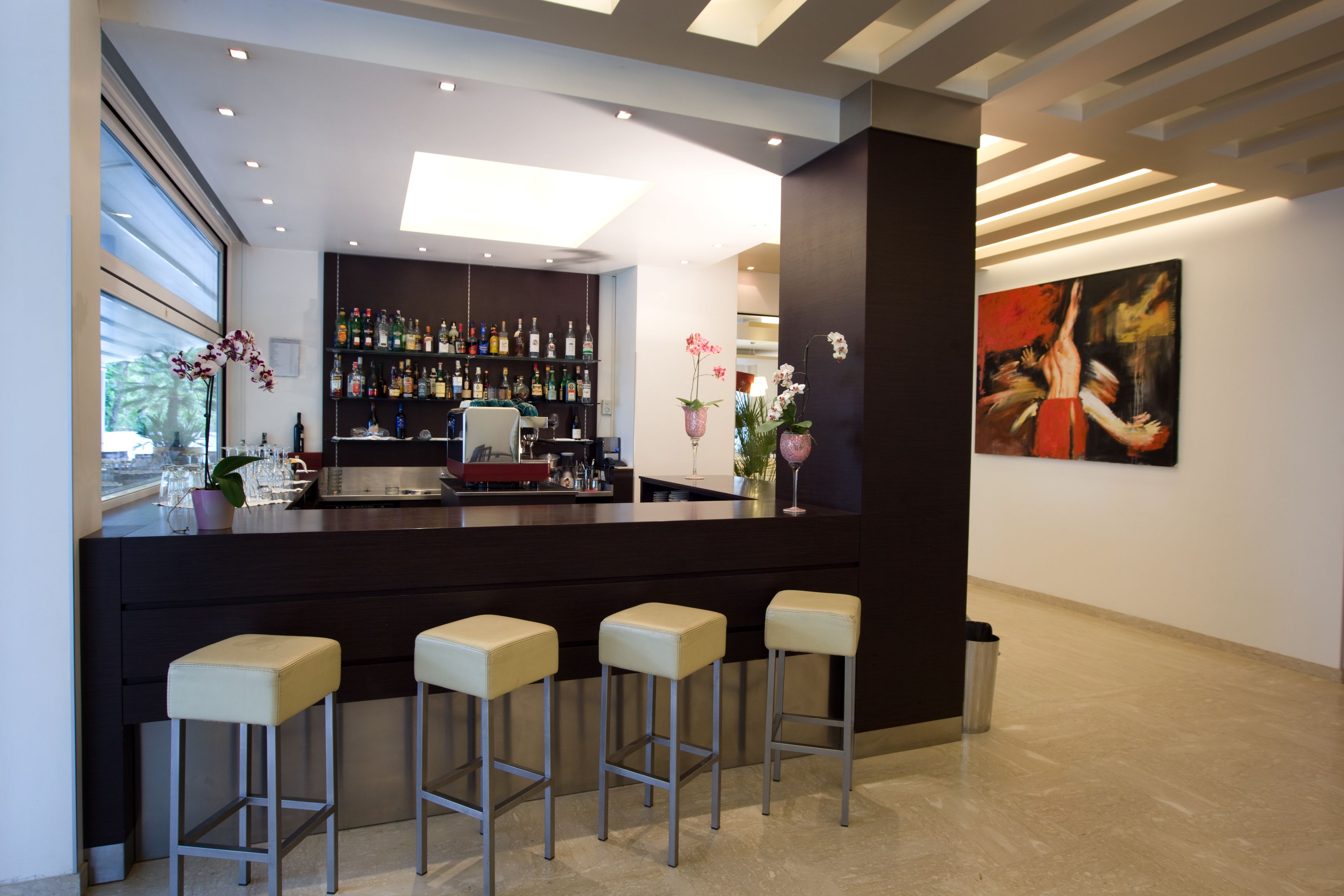 A modern bar area with a dark counter, three bar stools, a variety of bottles, and a large abstract painting on the wall.