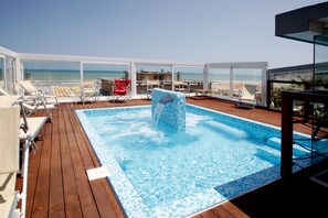 Una piscina en la azotea con vistas a la playa, rodeada de sillas de descanso y una mesa.