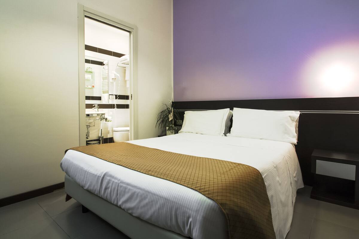 A neatly arranged hotel room with a large bed, a bedside table, and a bathroom visible through an open door.