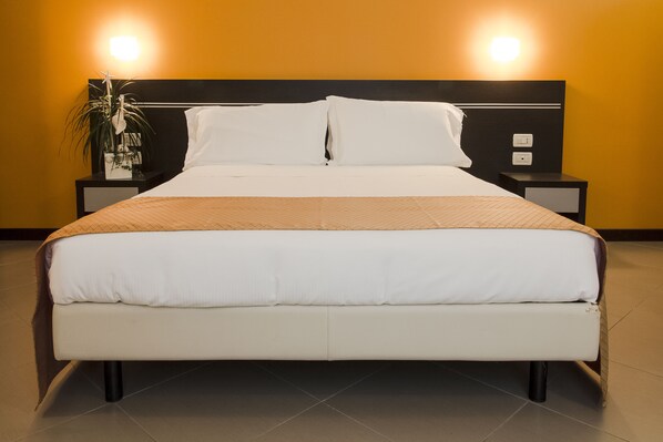 Una cama bien hecha con sábanas blancas y una manta marrón, flanqueada por dos mesitas de noche con lámparas y una planta en maceta.