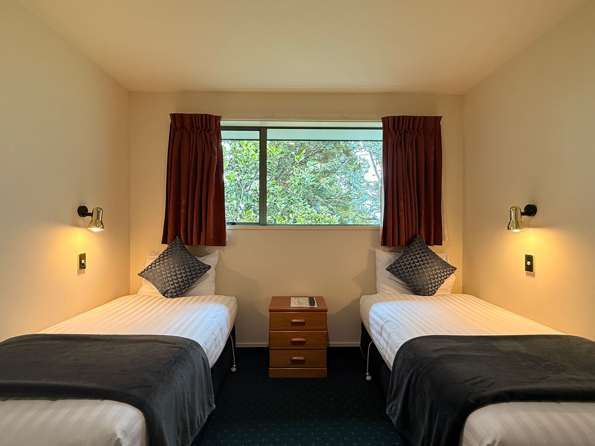 Habitación doble con dos camas, ventana con cortinas, lámparas de noche y un pequeño aparador de madera.
