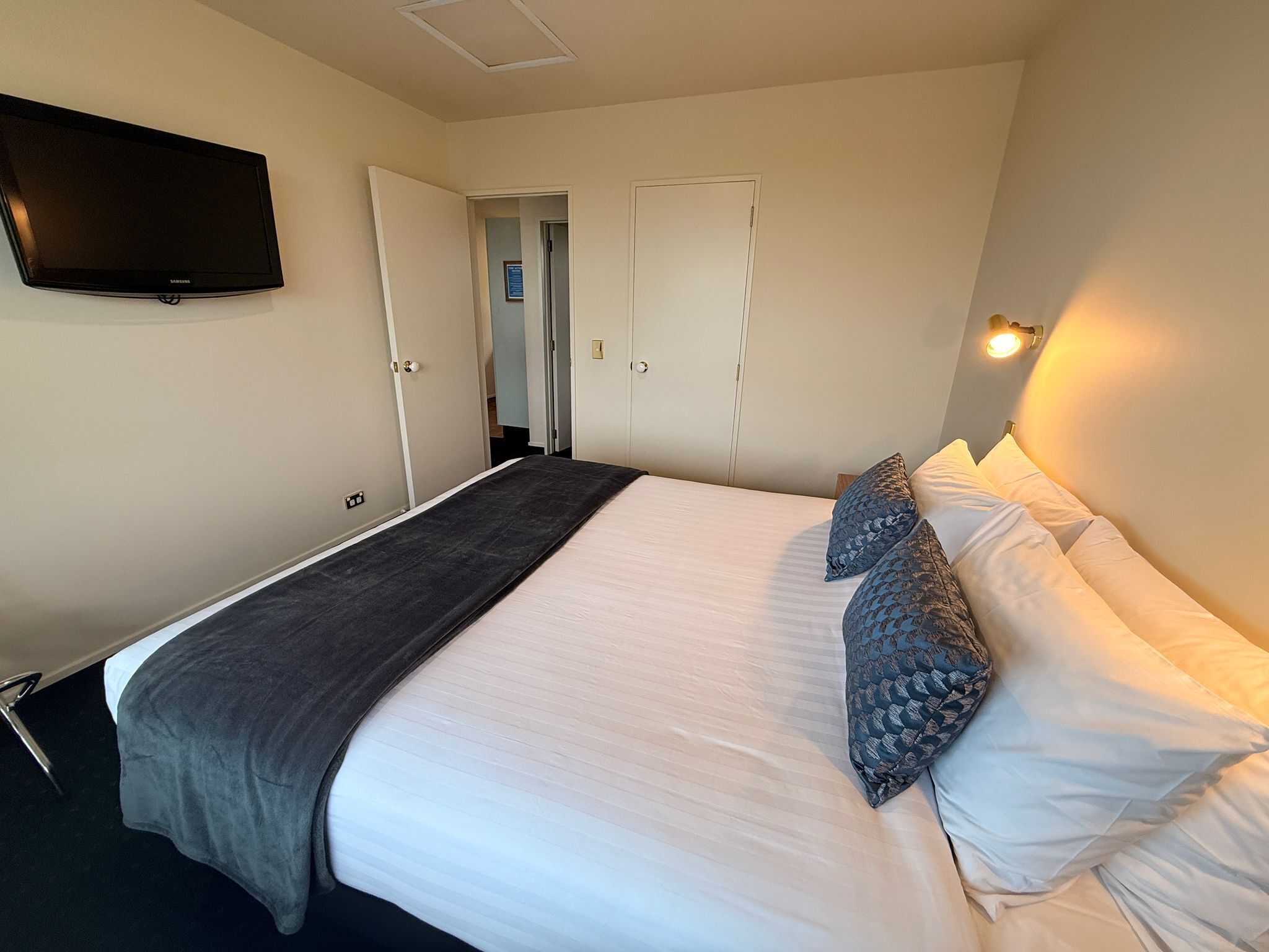 A neatly made bed with a grey blanket and patterned pillows, a flat-screen TV on the wall, and a door leading to another room.