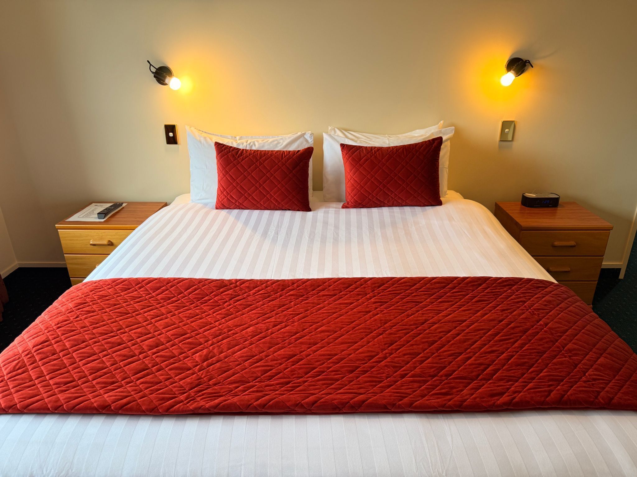Una cama bien hecha con colcha blanca y roja, dos almohadas rojas y dos lámparas fijadas a la pared.