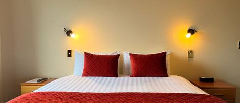 A hotel room with a large bed, two red pillows, a red blanket, and two bedside tables with lamps.