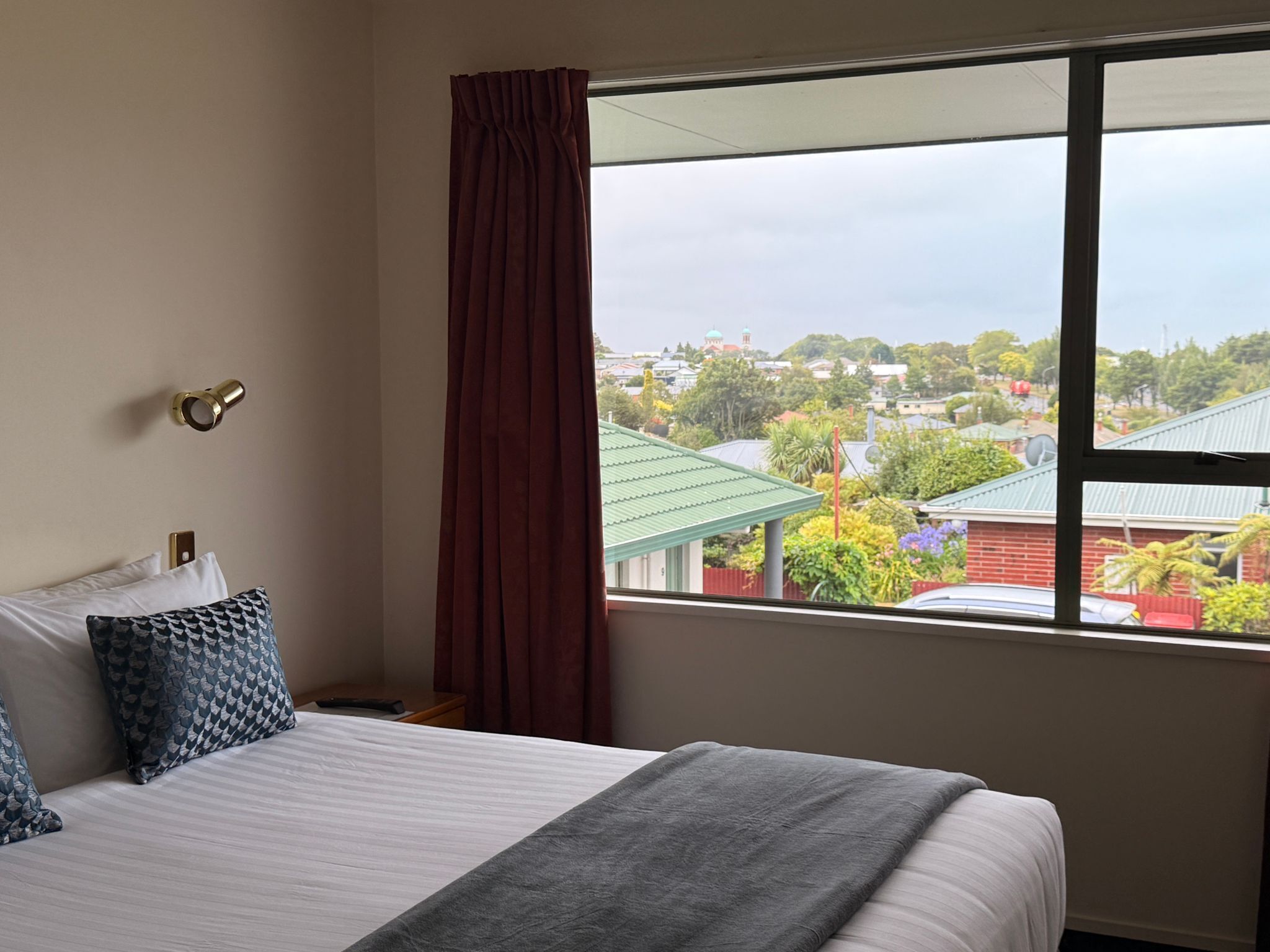 A hotel room with a bed, a window with a view of buildings and trees, and a curtain.