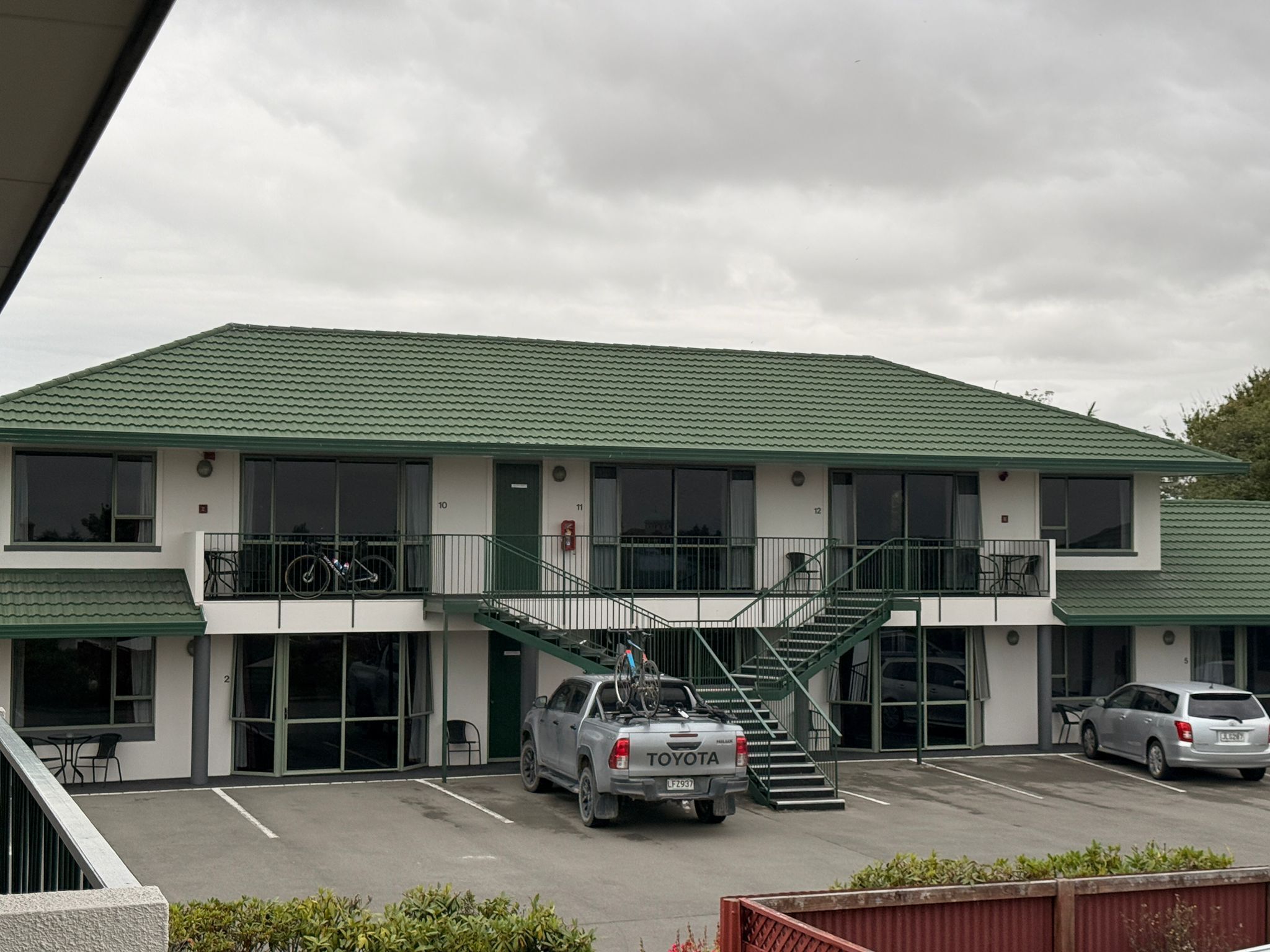 Un bâtiment à deux étages, doté d’un toit végétalisé et de murs blancs, avec un parking où sont garées plusieurs voitures, dont un pick-up Toyota.