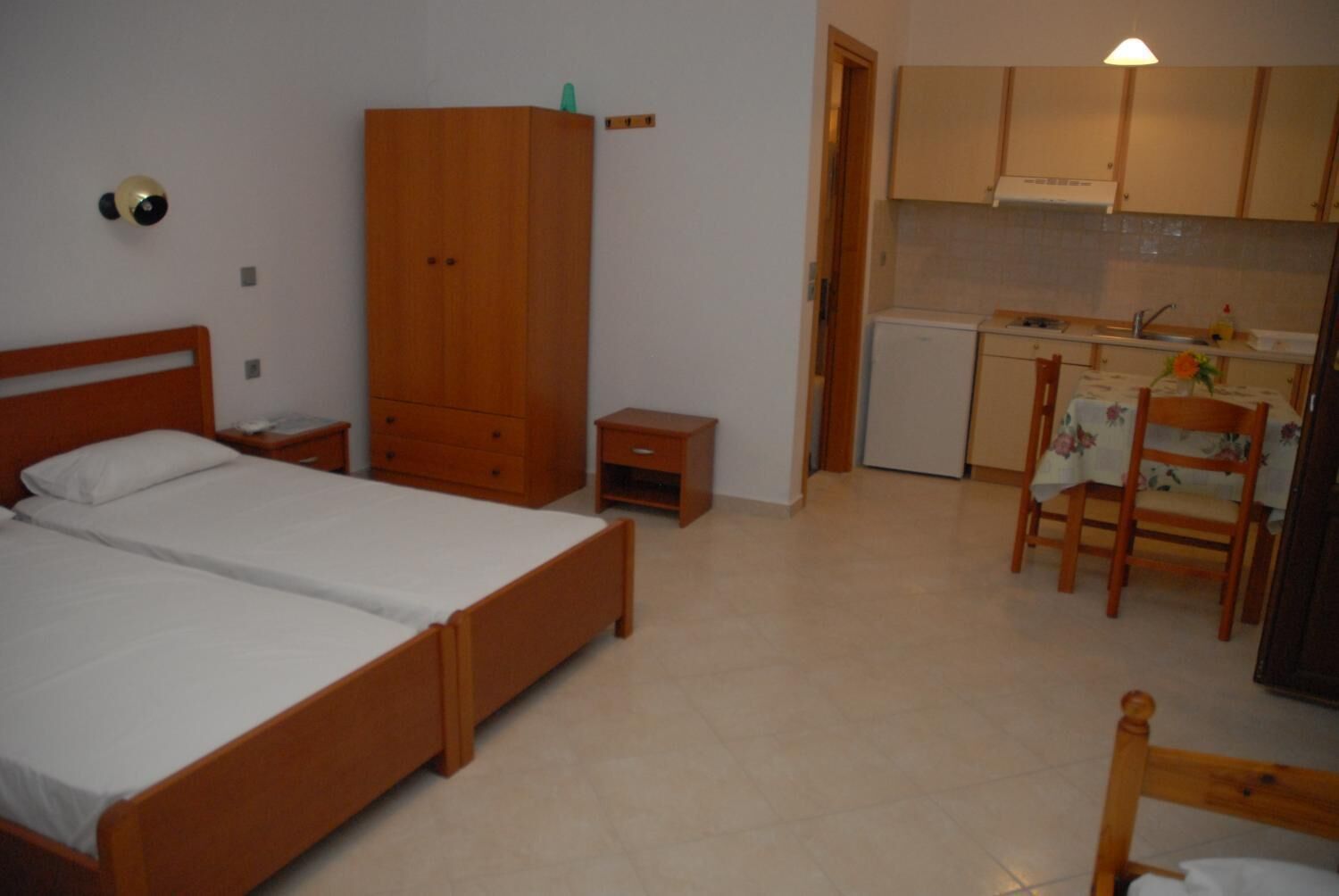 A bedroom with a double bed, a wooden wardrobe, a small bedside table, a nightstand, and a kitchenette with a sink, stove, and a chair with a table.