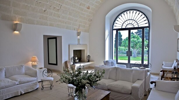 Un salon spacieux, meublé de blanc, avec une cheminée et une grande fenêtre en arc d’où l’on peut admirer la verdure.