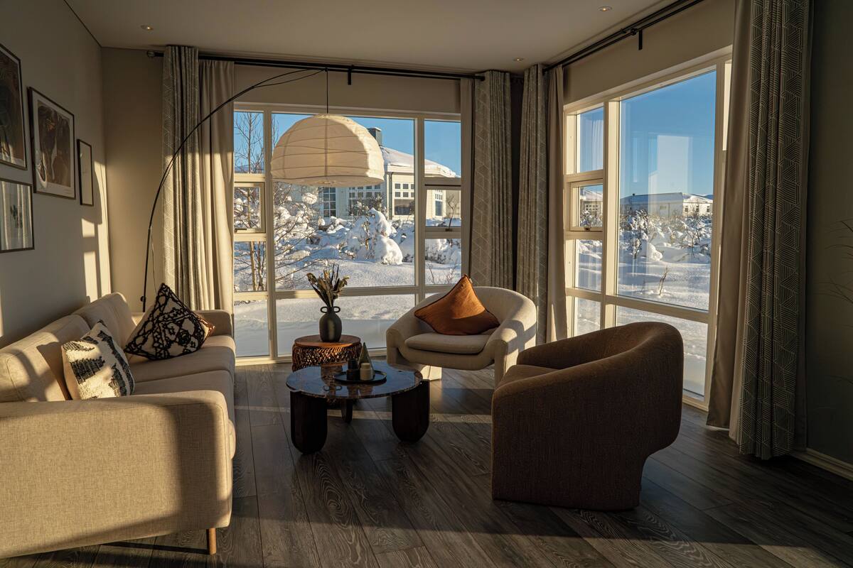 A modern living room with a sofa, armchair, and coffee table, large windows with a snowy view, and a hanging light fixture.