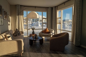Un salon moderne avec un canapé, un fauteuil et une table basse, de grandes fenêtres donnant sur un paysage enneigé et un luminaire suspendu.