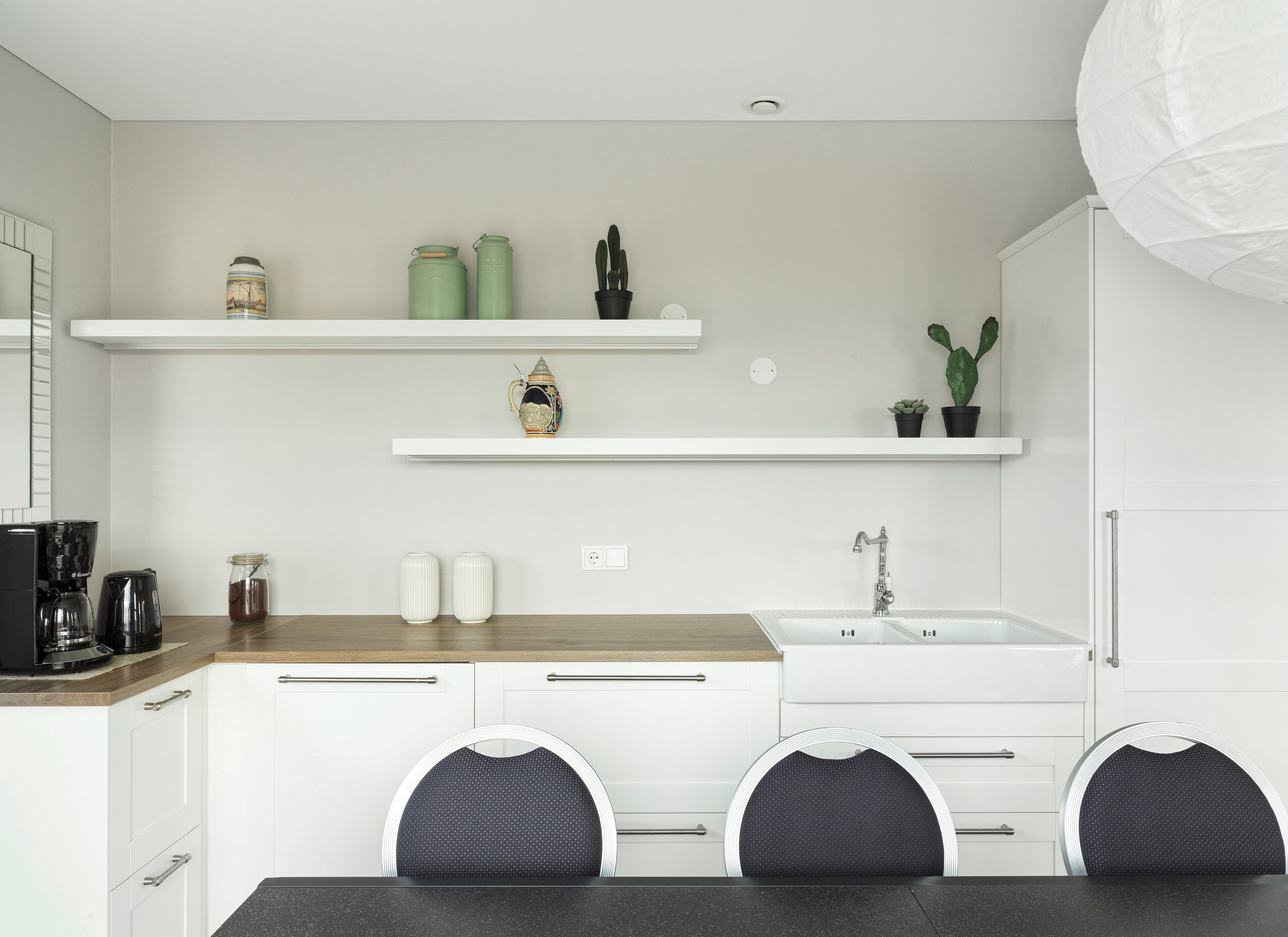 Une cuisine moderne avec des meubles blancs, un plan de travail en bois et un coin repas avec deux chaises. Au-dessus de l’évier, on trouve deux étagères : l’une est décorée d’objets et l’autre contient des plantes en pot.