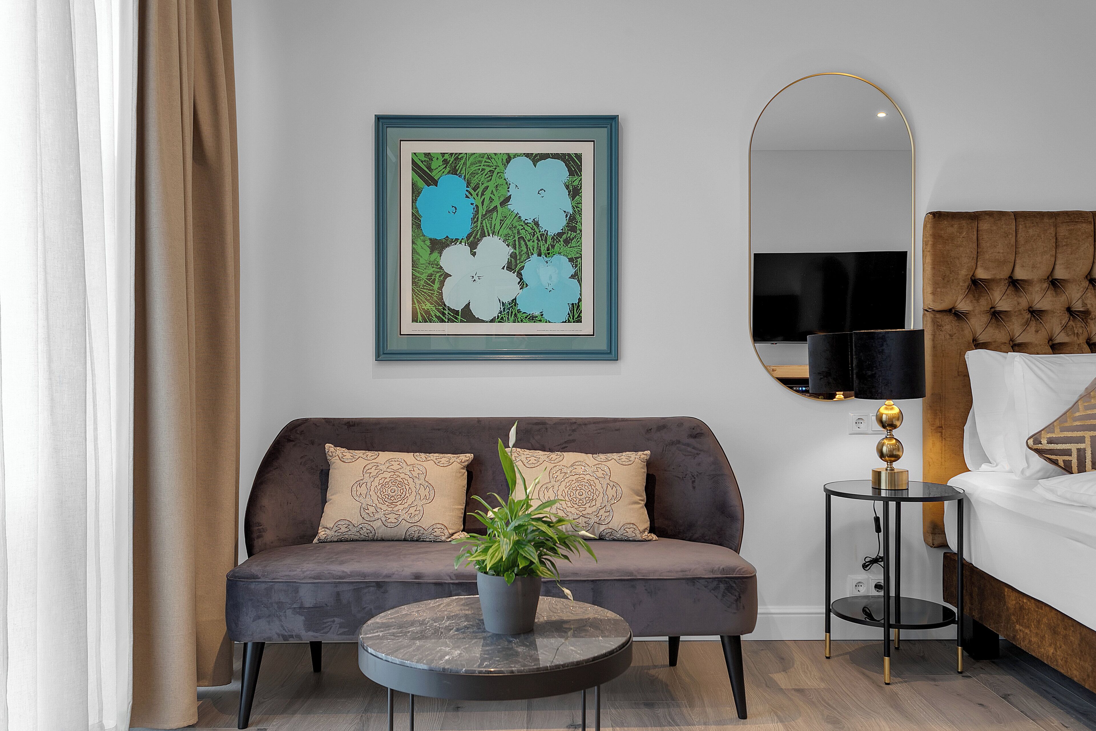A modern hotel room with a grey sofa, a round coffee table with a plant, a large mirror, and a bedside table with a lamp.