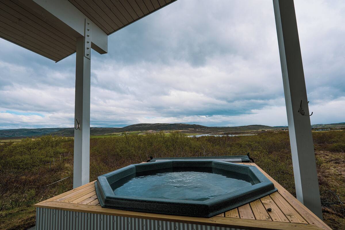 Un jacuzzi debajo de una terraza de madera, con vista a un paisaje cubierto de pasto y colinas al fondo.